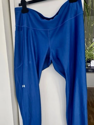 Blå leggings från Under Armour - Blå träningstights från Under Armour med hög midja och tight passform. Tillverkade i stretchigt funktionsmaterial som andas, perfekt för gym eller löpning. Diskret vit logga på benet. Använda men i väldigt bra skick, ej uttöjda 
