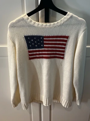 Vit stickad tröja med USA-flagga - Stickad tröja från SHEIN i vitt med en stor amerikansk flagga i blått, rött och vitt på bröstet. Tröjan har rund halsringning. Aldrig använd.