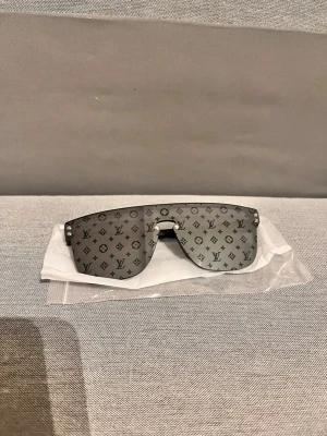 Luis Vitton monogram solglasögon - Snygga solglasögon från Luis Vitton med stora, rektangulära glas i silverton och ikoniskt monogrammönster över hela linsen. Bågarna är svarta och har diskret Luis Vitton-logga på sidorna. Perfekt accessoar för en trendig look.