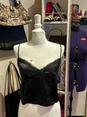 Svart satinlinne med spets - Elegant svart satinlinne från H&M med fin spetskant vid urringningen. Det mjuka satinliknande tyget ger en fin lyster och en feminin känsla. Tunna axelband ger en smickrande och stilren passform. Passar perfekt både till vardag och till mer uppklädda tillfällen.