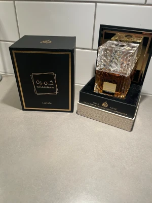Lattafa Khamrah 100ml - Exklusiv parfym från Lattafa, Khamrah, i en lyxig glasflaska med mönstrad design och guldiga detaljer. Flaskan har en fyrkantig form och innehållet är bärnstensfärgat. Levereras i elegant svart och guldig förpackning.
