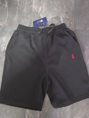 Svarta shorts från Polo Ralph Lauren - Svarta shorts från Polo Ralph Lauren med elastisk midja och snörning. Klassisk design med två sidofickor och den ikoniska röda logotypen broderad på vänster ben. Perfekta för en avslappnad och sportig stil.