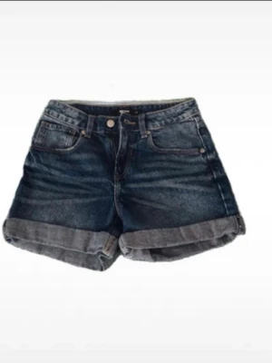 Mörkblå denimshorts med uppvik - Snygga mörkblå denimshorts med uppvikta benslut och klassisk femficksdesign. Shortsen har normal passform och midja, perfekta för sommardagar. Inga synliga loggor eller märken på utsidan.