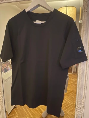 Svart t-shirt från C.P. Company - Stilren svart t-shirt från C.P. Company med klassisk rund hals och kort ärm. På ena ärmen finns en ikonisk detalj med logotypen i form av en lins. Perfekt för en clean och modern look.