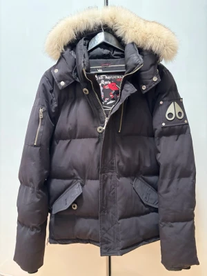 Svart dunjacka med pälskrage Moose Knuckles - Snygg svart dunjacka från Moose Knuckles med avtagbar huva och fluffig pälskant. Jackan har två stora fickor framtill, ärmficka med dragkedja och logga på ärmen. Perfekt för kalla vinterdagar och riktigt varm.