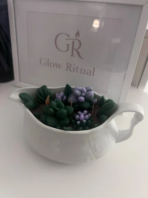 Dekorativ doftljus i kanna - Unik dekorativ ljuslykta från Glow Ritual i en vit porslinskanna. Ljuset är dekorerat med detaljer i form av gröna blad och lila blommor i vax, vilket ger ett naturinspirerat och konstnärligt intryck. Perfekt som inredningsdetalj eller present. HANDMADE