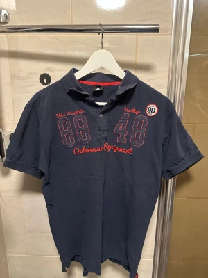 Mörkblå pikétröja med röd brodyr - Mörkblå pikétröja med kort ärm och krage, dekorerad med röd brodyr och siffror framtill. Tröjan har knappar vid halsen och sportig känsla. Perfekt för en avslappnad och stilren look. Den är lite sönder vid bröstet 