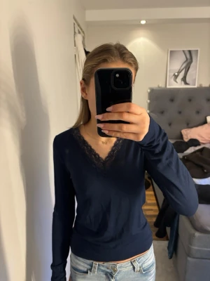 Marinblå långärmad topp med spets - Säljer en marinblå långärmad topp från Gina med v-ringning och dekorativ spets längs halsen. Toppens passform är normal och den är gjord i mjuk bomullsmix som känns skön mot huden. Perfekt att matcha med jeans eller kjol.