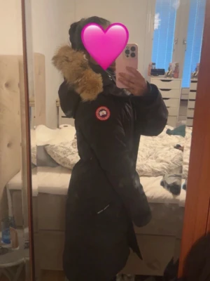 Svart parkas från Canada Goose - Snygg svart parkas från Canada Goose med klassisk logga på bröstet. Jackan har en stor huva med fluffig fuskpäls och praktiska fickor framtill. Perfekt för kalla dagar när du vill hålla dig varm och stilren.