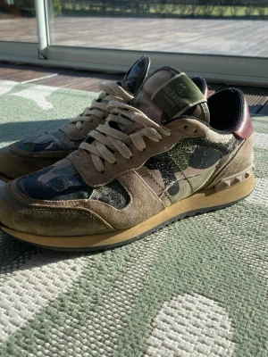 Valentino camo rockrunners med nitar - Snygga sneakers från Valentino med camo-mönster i grönt, beige och svart. Ovandel i mocka och textil, detaljer i läder och coola nitar på hälen. Skorna har beige snörning och en kontrasterande vinröd häl. Perfekta för dig som vill sticka ut med stil.