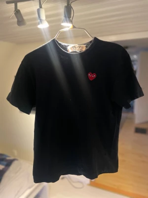 Svart Comme des Garçons t-shirt. Cdg t-shirt  - Svart t-shirt från Comme des Garçons Play med det ikoniska röda hjärtat broderat på bröstet. Klassisk passform med rund hals och korta ärmar. Perfekt för dig som gillar stilrena plagg med en twist. Finns två små hål (se bild 4), men som ni ser på bild 2 syns de inte mycket. Om ni har några funderingar så hör av er!🙂