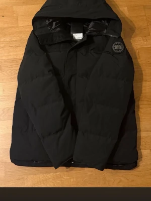 Svart pufferjacka från Canada Goose - Säljer en svart pufferjacka från Canada Goose med huva och diskret logotyp på ärmen. Jackan är tjockt vadderad och har en klassisk, boxig passform. Perfekt för kalla dagar när du vill hålla värmen och ändå se stilren ut.