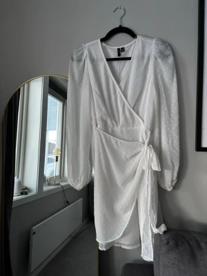 Vit omlottklänning med knyt - Snygg vit omlottklänning från Bubbleroom med knyt i midjan och diskret prickigt mönster. Klänningen har långa puffärmar, v-ringning och lätt volangkant nertill. Perfekt för sommarens alla tillfällen. Hör av dig vid frågor!🤍😊