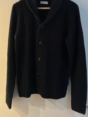 Svart kofta från Jack & Jones - Svart stickad kofta från Jack & Jones med grovt mönster och stora knappar framtill. Koftan har sjalkrage och ribbade muddar vid ärmslut och nederkant. Perfekt för lager-på-lager och en chill look. Storlek S 