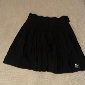 Svart plisserad kjol med knappdetalj - Säljer en svart plisserad kjol med bred linning och knappdetalj i midjan. Kjolen har klassiska veck och är tillverkad i ett mjukt tyg som ger ett snyggt fall. Perfekt för dig som gillar en stilren och tidlös look.