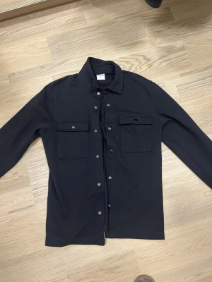 Svart overshirt - Svart overshirt från Selected Homme med bröstfickor. Skjortan har klassisk krage och knappar framtill. Perfekt att ha öppen eller stängd över en t-shirt för en avslappnad stil. 