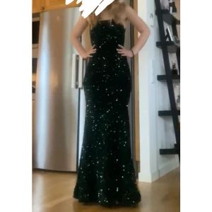 Mörkgrön glittrig balklänning - Säljer en mörkgrön balklänning med glittriga paljetter. Klänningen är kroppsydd och har öppen rygg med snörning. Klänningen är insydd i dragkedjan (se bild 3). Köpt för 900 i andra hand. Jag är en st S och är 167 cm lång (klackar på bilden)
