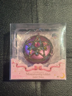 Sailor Moon Miniaturely Tablet - Säljer en Sailor Moon Miniaturely Tablet i originalförpackning. Tabletten är rund, metallicrosa med guldfärgade detaljer och färgade stenar i blått, rött och gult. Perfekt för dig som älskar anime och vill ha en unik accessoar till din samling.