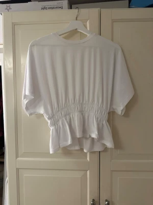 Vit topp med smock från 157 - Vit topp från 157 med smockad midja och volangkant nedtill. Modellen har korta ärmar och rund halsringning. Tillverkad i mjukt bomullstyg som ger en avslappnad och luftig känsla. Perfekt för dig som gillar en enkel men ändå söt stil.