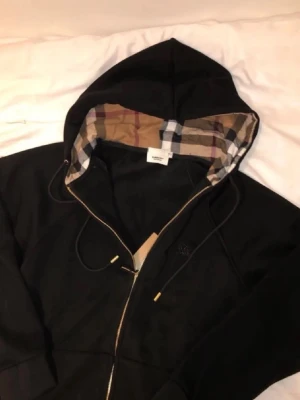 Svart hoodie från Burberry - Säljer en svart hoodie från Burberry med dragkedja och klassiskt rutigt foder i huvan. Hoodien har guldfärgade detaljer på snörena och muddar vid ärmslut och nederkant. Perfekt för dig som gillar stilrena plagg med exklusiva detaljer. S/ liten m 