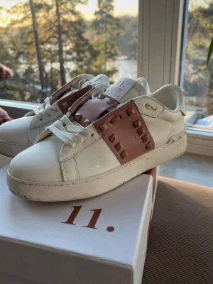 Valentino Rockstud Untitled sneakers - Snygga vita sneakers från Valentino med ljusrosa läderdetaljer och ikoniska nitar över snörningen. Modellen heter Rockstud Untitled och har en chunky sula och klassisk låg profil. Perfekta för dig som vill sticka ut med en lyxig touch.