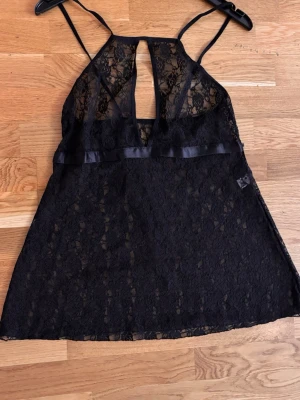 Svart spetsnattlinne med smala axelband - Snyggt svart nattlinne i genombruten spets med smala axelband och dekorativ öppning framtill. Nattlinnet har en lätt A-linjeform och ett satinband under bysten för extra detalj. Perfekt för en lyxig och bekväm känsla.