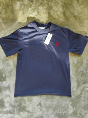 Mörkblå t-shirt från Ami Paris - Snygg mörkblå t-shirt från Ami Paris med röd broderad logga på bröstet. Klassisk passform med korta ärmar och rund halsringning. Tillverkad i mjuk bomull som känns skön mot huden. Perfekt för en clean och stilren look.