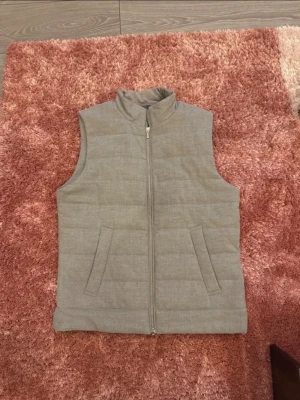 Ljusgrå dunväst med dragkedja - Stilren ljusgrå dunväst med hög krage och dragkedja framtill. Västen har två snedställda fickor och quiltad design för extra värme. Passar perfekt över en hoodie eller tröja för en avslappnad look.