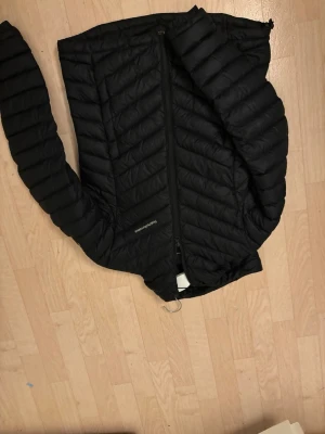 Svart dunjacka från Peak Performance - Snygg svart dunjacka från Peak Performance med quiltat mönster och dragkedja framtill. Jackan har en stilren design och är perfekt för kalla vinterdagar. Liten logga på bröstet och lätt vaddering som håller dig varm utan att kännas klumpig.vet inte varför bilderna blev upp å ner!