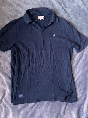 Marinblå pikétröja från Morris - Klassisk marinblå pikétröja från Morris med liten broderad logga på bröstet. Tröjan har korta ärmar, krage och knappar vid halsen. Perfekt till jeans eller shorts för en avslappnad men stilren look. En strykning kan behövas men har inte orkat göra de derav priset.