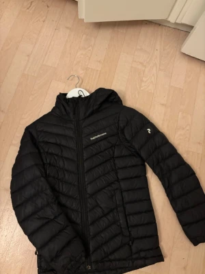 Svart dunjacka från Peak Performance - Snygg svart dunjacka från Peak Performance med huva och dragkedja framtill. Jackan har två sidofickor med dragkedja och diskret logga på bröstet samt ärmen. Perfekt för kalla vinterdagar och riktigt lätt att bära.