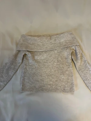 Beige grå  off shoulder stickad tröja - Trendig beige stickad tröja med off shoulder-design och bred krage. Tröjan har ribbstickat mönster och långa ärmar.