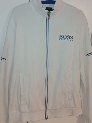 Vit ziptröja från Hugo Boss (L/XL/ - Passar även L. Snygg vit ziptröja från Hugo Boss med blå logga på bröstet och blå detaljer längs dragkedjan och ärmarna. Tröjan har hög krage, två fickor framtill och ribbade muddar. Perfekt för en clean och sportig stil.