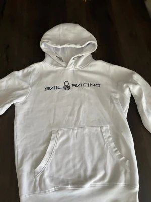 Vit hoodie från Sail Racing - Snygg vit hoodie från Sail Racing med svart logga tryckt över bröstet. Klassisk känguruficka framtill och stor huva. Tillverkad i mjukt material, perfekt för en avslappnad och sportig stil.
