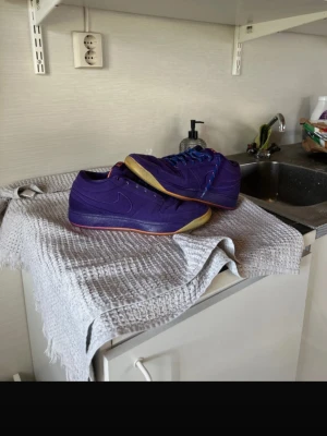 D.Book 1 - Säljer ett par lila Nike sneakers med klassisk låg profil och diskret swoosh på sidan. Skorna har en kontrasterande gummisula i beige och röda detaljer på hälen. Ena skon har blå skosnören och den andra har röda, vilket ger en unik look. Perfekta för dig som vill sticka ut.