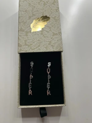 Silvriga örhängen med bokstäver från IOAKU  - Säljer ett par statement-örhängen i silverfärgad metall där bokstäverna bildar ordet 'SUPER' på varje örhänge. Hängande design med varje bokstav länkad i rad. Perfekta för dig som vill sticka ut och addera en cool detalj till din stil. ⚠️kostar 600 kr, ALDIG ANVÄNDA⚠️