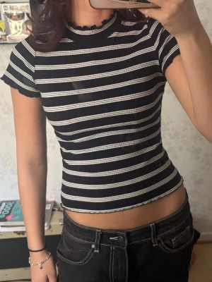 Svartvit ribbad randig topp - Snygg svart ribbad topp med vita ränder och vågiga kanter vid hals, ärmslut och nederkant. Kortärmad och figurnära modell som ger en trendig och ungdomlig look. Perfekt att matcha med jeans eller kjol.