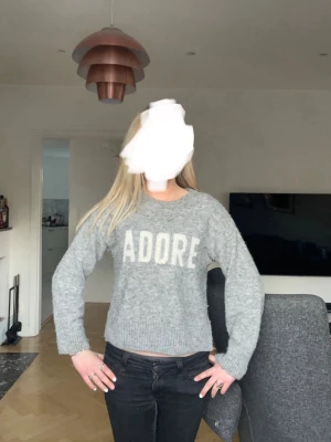 Grå stickad tröja med text - Mysig grå stickad tröja med vit text 'ADORE' framtill. Tröjan har rund halsringning, långa ärmar och en något kortare, boxig passform. Perfekt för att skapa en avslappnad och trendig look.