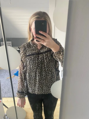 Leopardmönstrad blus med volang - Snygg leopardmönstrad blus med volangdetaljer och svarta spetsband över bröstet. Blusen har långa ärmar med resår vid ärmslut och en lös, bekväm passform. Storlek 158/164