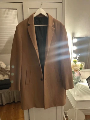 Klassisk kamelfärgad ullkappa - Stilren kamelfärgad kappa med rak passform och svarta knappar. Kappan har två fickor framtill, klassisk krage och är helfodrad med svart innerfoder. Perfekt för dig som vill ha en tidlös och snygg look till hösten och vintern.