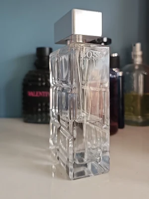 Hugo Boss glasflaska parfym - Stilren parfymflaska från Hugo Boss i transparent glas med geometriskt mönster och en stor, silverfärgad fyrkantig kork. Flaskan har en modern och lyxig känsla, perfekt för dig som gillar eleganta detaljer på parfymhyllan.