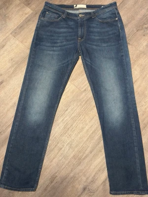 Blå raka jeans med slitningar - Snygga mörkblå jeans från Dressmann med lätt tvättad effekt. Klassisk 5-ficksmodell i regular fit med skön stretch för bra komfort. ✔ Fint skick – normalt slitage ✔ Stretchig denim (94% bomull, 5% polyester, 1% elastan) ✔ Regular fit (rak passform) ✔ Passar till både vardag och lite mer uppklätt Tvättade och redo att användas! 📦 Skickas snabbt 💬 Hör av dig vid frågor!