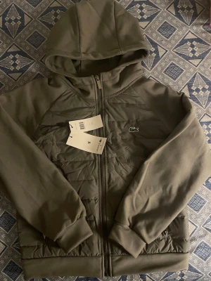 Olivgrön hoodie från Lacoste - Säljer en olivgrön hoodie från Lacoste med dragkedja och huva. Framsidan har det klassiska krokodilmärket broderat. Materialet är en mix av quiltad front och mjuka ärmar, vilket ger en sportig och modern look. Perfekt för dig som gillar streetwear och stilrena detaljer.