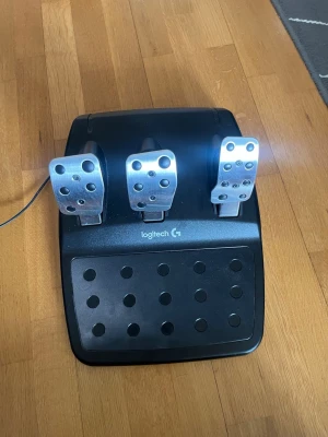 Logitech G29 ratt och pedaler - Logitech G29 racingratt och pedalset för PlayStation och PC. Ratten är klädd i svart läder med blå detaljer och har flera knappar samt en röd vridreglage. Pedalerna är i metall och sitter på en robust svart plastbas. Perfekt för racing- och bilspel.