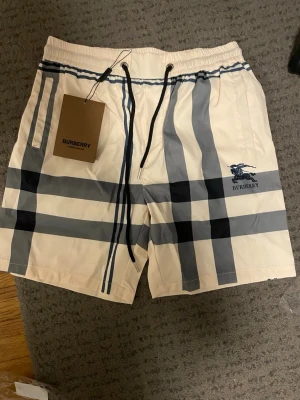 Rutig Burberry shorts med snörning - Säljer ett par snygga Burberry shorts i klassiskt beige med blå och svarta rutor. Shortsen har elastisk midja med svart snörning och sidofickor. Ikoniskt Burberry-märke broderat framtill. Perfekta för sommaren och riktigt trendiga. Pris kan diskuteras