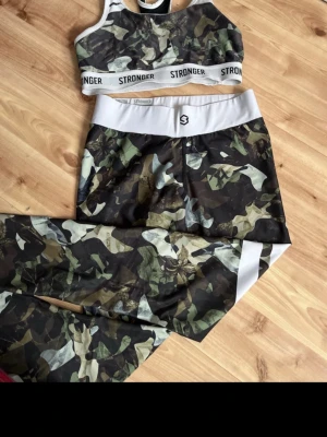 Camo träningsset från Stronger - Snyggt träningsset från Stronger med camouflagemönster i grönt, svart och vitt. Setet består av en sport-bh med bred resår och matchande tights med hög midja och vita detaljer. Perfekt för gymmet eller löprundan.
