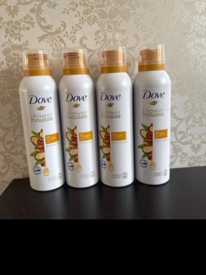 Dove Shower Mousse Arganolja - Fyra flaskor Dove shower mousse med arganolja. Flaskorna är vita med guldiga lock och detaljer, och har en illustration av argannötter på framsidan. Moussen är avsedd för dusch och ger en lyxig känsla med mjukgörande egenskaper.