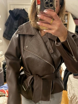 Brun oversized skinnjacka med bälte - Säljer en brun oversized skinnjacka med bred krage, snedställd dragkedja och bälte i midjan. Jackan har coola detaljer som dragkedjor vid ärmsluten och stora slag. Använt en gång, pris kan diskuteras vid snabb affär