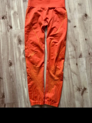 Orange sportleggings med mönster - Snygga orange sportleggings med högt midjeband och tunna gula linjer som skapar ett sportigt mönster längs benen. Tillverkade i stretchigt funktionsmaterial som passar perfekt för träning eller löpning. Tight passform och sömlös design för extra komfort.