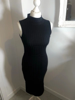 Svart ribbad ärmlös klänning - Snygg svart ribbad klänning med hög krage och ärmlös design. Klänningen är figurnära och har en längre modell som ger en stilren och modern look. Perfekt för dig som gillar minimalistisk stil och vill ha något enkelt men ändå trendigt.